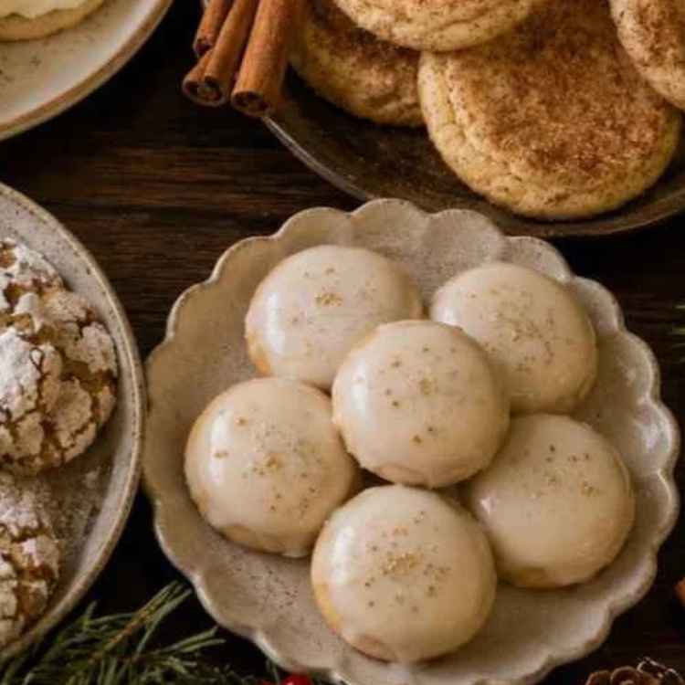 A plate of six round cookies with white icing, surrounded by other cookies and cinnamon sticks on a wooden table.