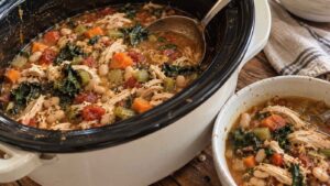 Slow cooker filled with chicken, white bean, and kale soup, next to a bowl of this healthy crockpot recipe.